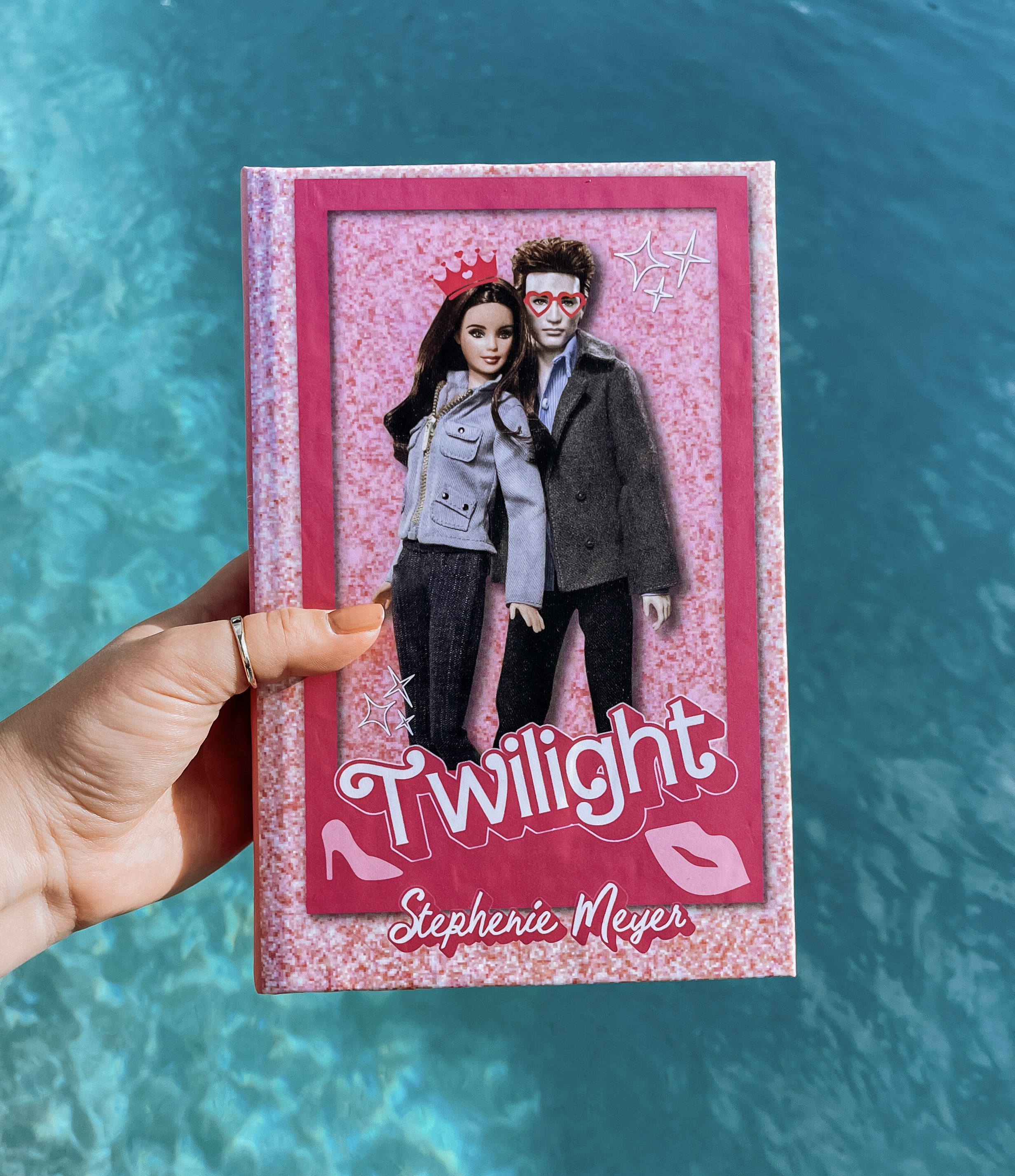 Cover of Twilight pink rebind, hand is holding the book with a blue water background